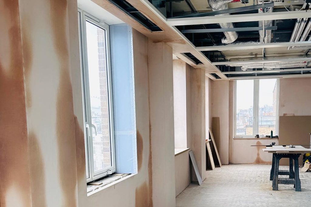 Commercial office fit-out in progress: freshly plastered partition walls and window reveals, with an exposed-services ceiling zone and a city skyline beyond.